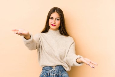 Beyaz tenli genç bir kadın bej arka planda izole edilmiş, kollarıyla ölçek yapıyor, mutlu ve kendinden emin hissediyor..