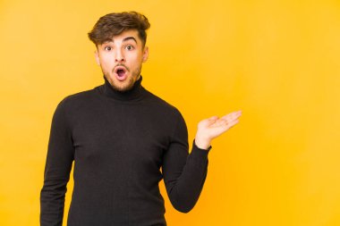 Sarı arka planda izole edilmiş genç bir Arap, avuç içi fotokopi alanını tutmaktan etkilenmiş..