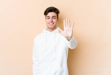 Beyaz tenli genç adam bej arka planda izole edilmiş. Gülümsüyor. Beş numarayı parmaklarıyla gösteriyor..