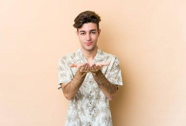 Beyaz tenli genç adam bej arka planda izole edilmiş. Elinde avuç içi olan bir şeyle kameraya sunuyor..