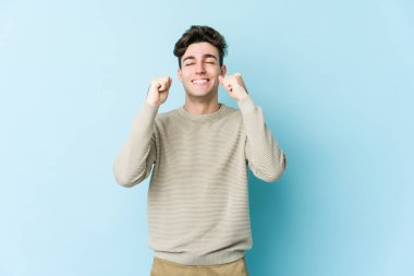 Mavi arka planda izole edilmiş genç beyaz bir adam zaferi, tutkuyu ve coşkuyu kutluyor, mutlu bir ifade..