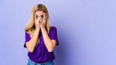 Mor arka planda izole edilmiş genç sarışın kadın kameraya parmaklarıyla göz kırpıyor, yüzünü kapatmaktan utanıyor..