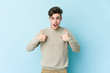 Mavi arka planda izole edilmiş genç beyaz adam parmağını göstererek şaşırdı, geniş bir gülümsemeyle..