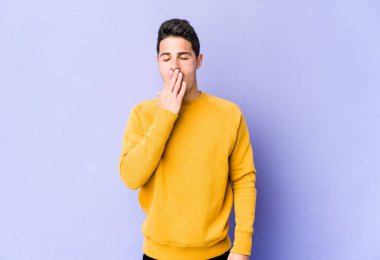 Mor arka planda izole edilmiş genç beyaz adam esniyor. Ağzı eliyle kapatan yorgun bir hareket sergiliyor..