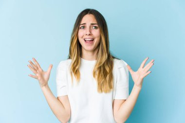 Mavi arka planda izole edilmiş genç beyaz kadın gökyüzüne haykırıyor, yukarı bakıyor, hayal kırıklığına uğramış..