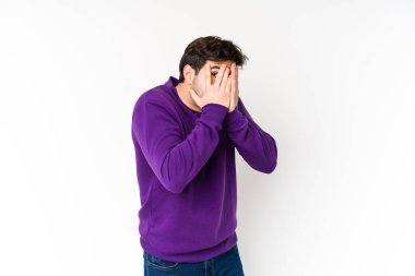 Beyaz arka planda izole edilmiş genç adam parmaklarının arasından korkmuş ve gergin bir şekilde göz kırpıyor..