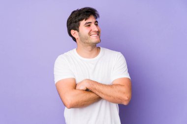 Mor arka planda izole olmuş genç adam kollarını kavuşturarak gülümsüyor..
