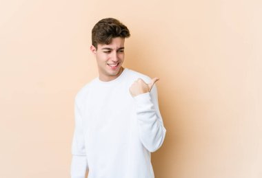 Beyaz tenli genç adam bej arka planda izole edilmiş, başparmak uzakta, gülüyor ve kaygısız..