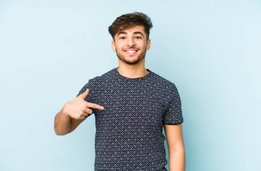 Mavi arka planda yaşayan genç bir Arap adam gömlek fotokopi alanını elle gösteriyor, gururlu ve kendinden emin.