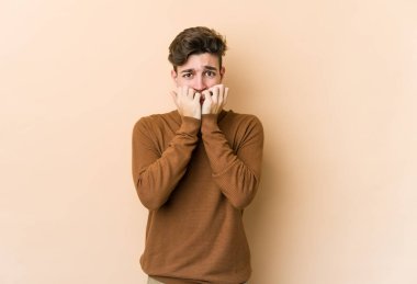 Beyazlı genç bir adam bej arka planda izole edilmiş tırnak yiyor, gergin ve endişeli..