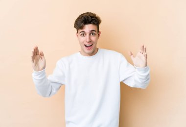 Beyaz tenli genç adam bej arka planda izole edilmiş. Hoş bir sürpriz bekliyor. Heyecanlı ve el kaldırıyor..