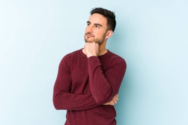 Mavi arka planda izole edilmiş genç beyaz adam şüpheli ve şüpheci bir ifadeyle yana bakıyor..