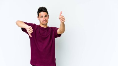 Beyaz arka planda izole edilmiş genç beyaz bir adam baş parmaklarını aşağı ve yukarı kaldırıyor, zor seçim konsepti