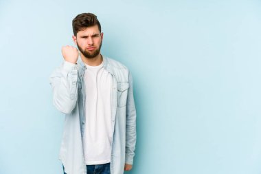 Mavi arka planda izole edilmiş genç beyaz adam yumruğunu kameraya gösteriyor, agresif yüz ifadesi var..