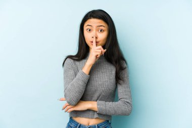 Genç Çinli bir kadın mavi arka planda izole edilmiş bir şekilde ya sır saklıyor ya da sessizlik istiyor..