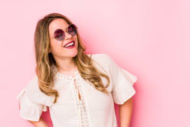 Gözlüklü genç beyaz kadın pembe arka planda izole edilmiş rahat ve mutlu bir şekilde gülüyor, boynunu uzatmış dişlerini gösteriyor..