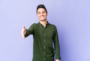 Mor arka planda izole edilmiş genç beyaz adam selamlama jesti olarak kameraya elini uzatıyor..
