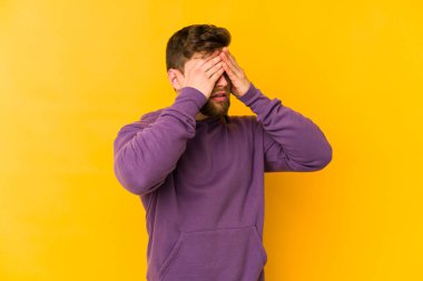 Sarı arka planda izole edilmiş genç beyaz adam elleriyle gözlerini kapatmaktan korkuyor..