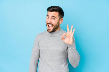 Mavi arka planda genç beyaz bir adam bir göz kırptı ve elleriyle iyi bir jest yaptı..