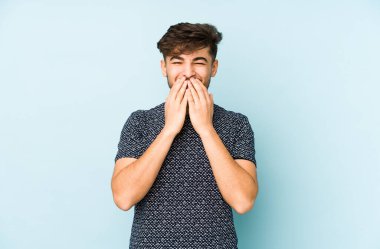 Genç Arap bir adam mavi bir arka planda izole edilmiş. Bir şeye gülüyor, elleriyle ağzını kapatıyor..