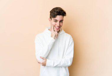 Beyaz tenli genç adam bej arka planda izole edilmiş mutlu ve kendinden emin gülümsüyor, elleriyle çenesine dokunuyor..