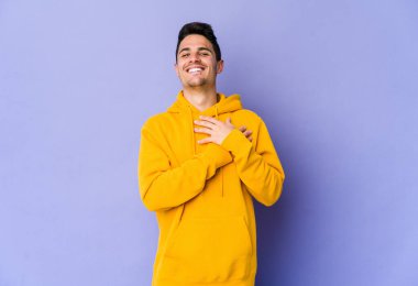 Mor arka planda izole olmuş genç beyaz adam gülüyor, ellerini yüreğine koyuyor, mutluluk kavramını koruyor..