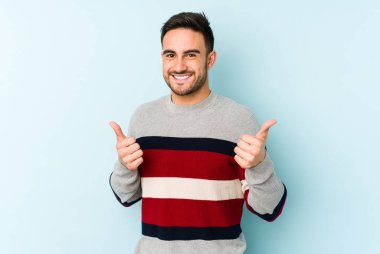Kafkasyalı genç adam mavi arka planda izole edilmiş. Baş parmağını kaldırıyor, gülümsüyor ve kendinden emin..