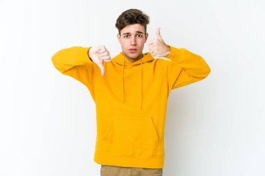 Beyaz arka planda izole edilmiş genç beyaz bir adam baş parmaklarını aşağı ve yukarı kaldırıyor, zor seçim konsepti