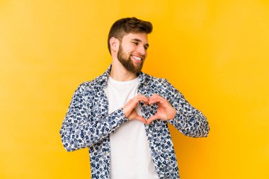 Sarı arka planda izole edilmiş genç beyaz adam gülümsüyor ve elleriyle kalp şeklini gösteriyor..