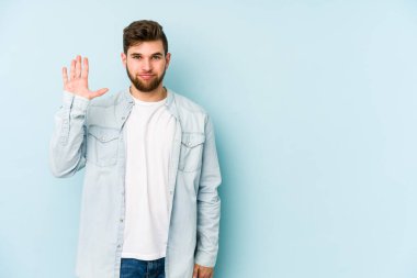 Mavi arka planda izole edilmiş genç beyaz adam gülümseyerek beş numarayı parmaklarıyla gösteriyor..