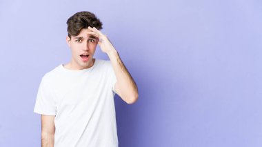 Mor arka planda izole edilmiş genç beyaz adam uzaklara bakıyor ve elini alnına koyuyor..