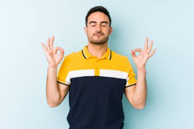 Mavi arka planda izole edilmiş genç beyaz adam yorucu bir günün ardından rahatlıyor. Yoga yapıyor..