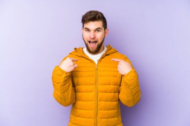 Mor arka planda izole edilmiş genç adam parmağını göstererek şaşırdı, geniş bir gülümsemeyle..