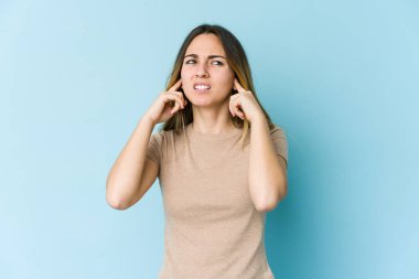 Genç beyaz kadın mavi arka planda izole edilmiş parmaklarıyla kulaklarını kapatıyor, gürültülü bir ortam yüzünden stresli ve çaresiz..