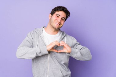 Mor arka planda izole edilmiş genç adam gülümsüyor ve elleriyle kalp şeklini gösteriyor..
