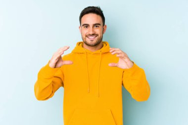 Kafkasyalı genç adam mavi arka planda izole edilmiş. Elinde avuç içi olan bir şey var ve kameraya sunuyor..