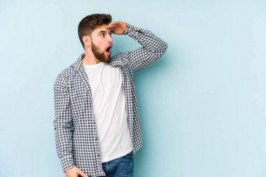 Kafkasyalı genç adam mavi arka planda izole edilmiş, uzaklara bakıyor ve elini alnına koyuyor..