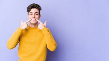 Mor arka planda izole edilmiş genç beyaz adam gülümsüyor, parmaklarını ağıza doğrultuyor..