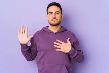 Mor arka planda izole edilmiş genç beyaz adam yemin ediyor, elini göğsüne koyuyor..