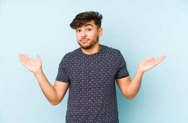 Genç Arap bir adam mavi bir arka planda izole edilmiş sorgulama sırasında omuz silkiyor ve kuşkulanıyor..