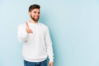 Mavi arka planda izole edilmiş genç beyaz adam selamlama jesti olarak kameraya elini uzatıyor..