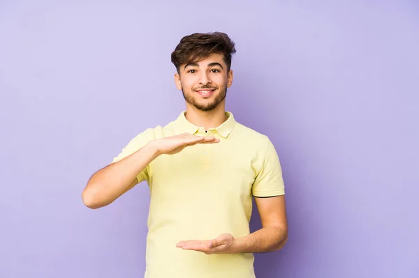 両手で何かを持っている紫色の背景に隔離された若いアラビア人の男 製品のプレゼンテーション — ストック写真