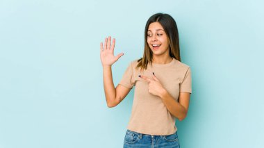 Mavi arka planda izole edilmiş genç bir kadın gülümseyerek beş numarayı parmaklarıyla gösteriyor..