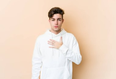 Beyaz tenli genç adam bej arka planda izole edilmiş yemin ediyor, elini göğsüne koyuyor..