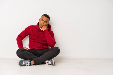 Yerde yalnız başına oturan genç latin adam üzgün ve düşünceli, fotokopi odasına bakıyor..