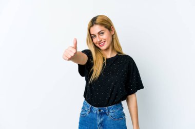 Beyaz arka planda izole edilmiş genç sarışın kadın gülümsüyor ve başparmağını kaldırıyor.