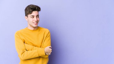 Mor arka planda izole edilmiş genç beyaz adam kollarını kavuşturarak gülümsüyor..