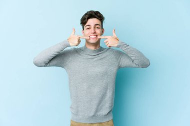 Mavi arka planda izole edilmiş genç beyaz adam gülümsüyor, parmaklarını ağıza doğrultuyor..