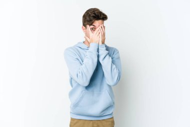 Beyaz arka planda izole edilmiş genç beyaz bir adam parmaklarının arasından korkmuş ve gergin bir şekilde göz kırpıyor..