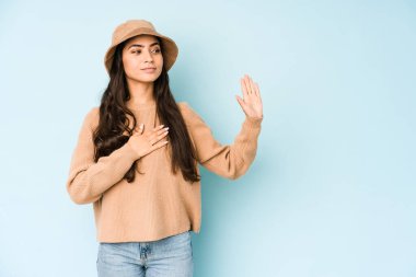 Mavi arka planda izole edilmiş bir şapka takan genç Hintli kadın yemin ediyor, elini göğsüne koyuyor..